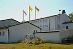 Ein großer Gebäudekomplex umgeben von Rasen. Im Vordergrund drei Fahnenmasten mit der Thailänischen, Deutschen und Buddhistischen Flagge.