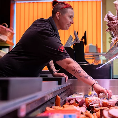 Eine Mitarbeiterin der Metzgerei greift hinter einer gut gefüllten Verkaufstheke mit verschiedenen Wurst- und Fleischwaren nach einem Produkt.