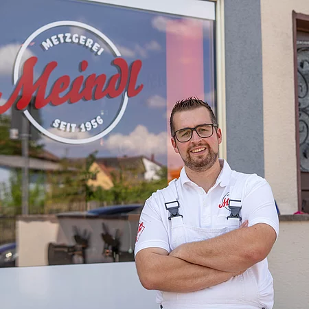 Ein Mitarbeiter der Metzgerei steht mit verschränkten Armen vor dem Geschäft; hinter ihm ist im Schaufenster das große Logo „Metzgerei Meindl – Seit 1956“ zu sehen.